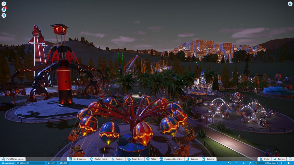 Planet Coaster (Windows) screenshot: As the sun sets on the amusement park, all the rides start to light up